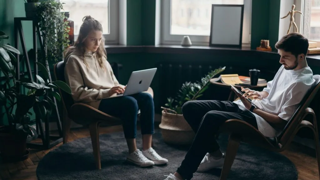 Woman and man sitting in office chairs facing each other, woman using laptop, man writing on clipboard, surrounded by plants and windows