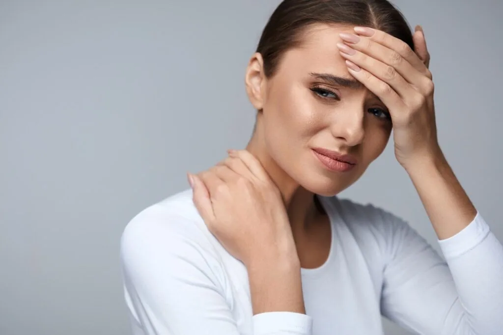 Woman in white shirt holding her forehead and neck, showing signs of pain or discomfort