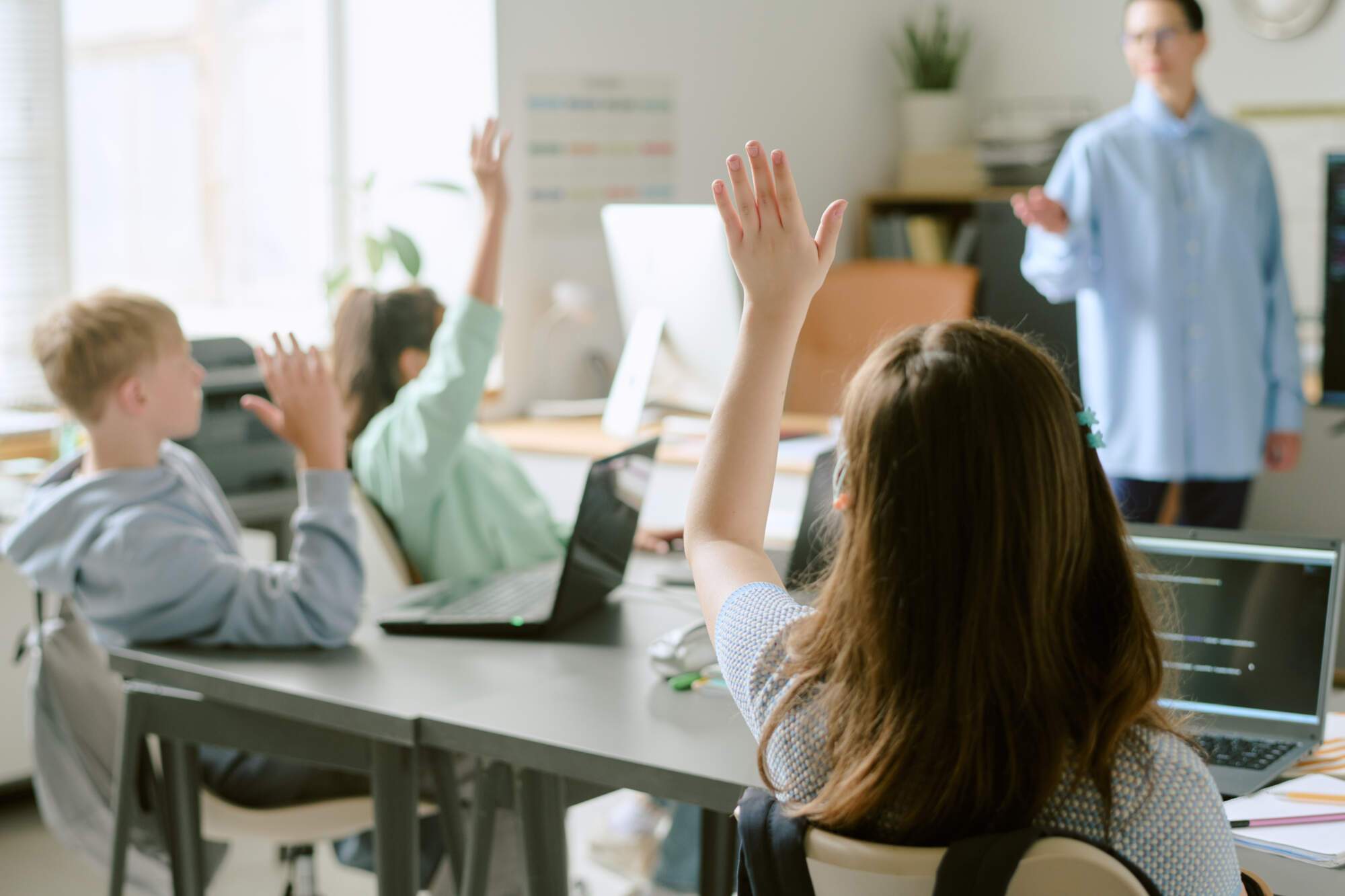 Students sitting at desks with laptops, raising their hands in a classroom with a teacher standing in the background.