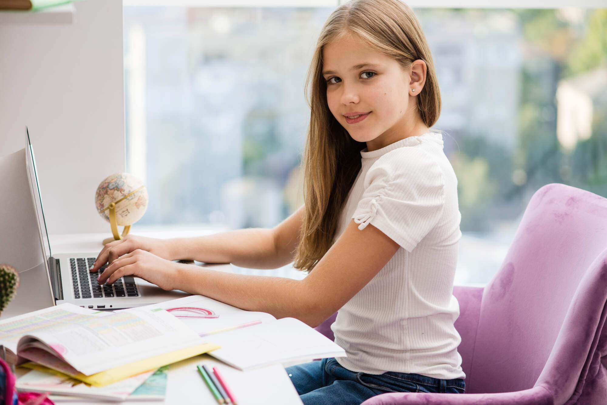 Young girl with long blonde hair sitting at a desk with a laptop, globe, and school supplies, looking over her shoulder at the camera with a slight smile