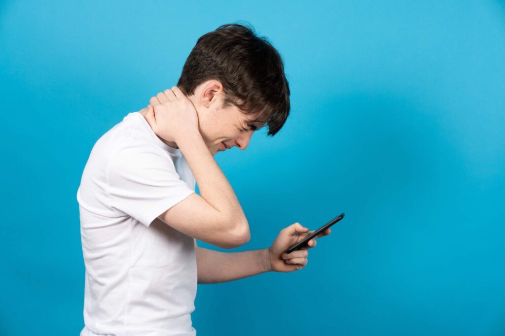 Teenage boy in white shirt holding smartphone with head bent forward, touching neck in pain against blue background